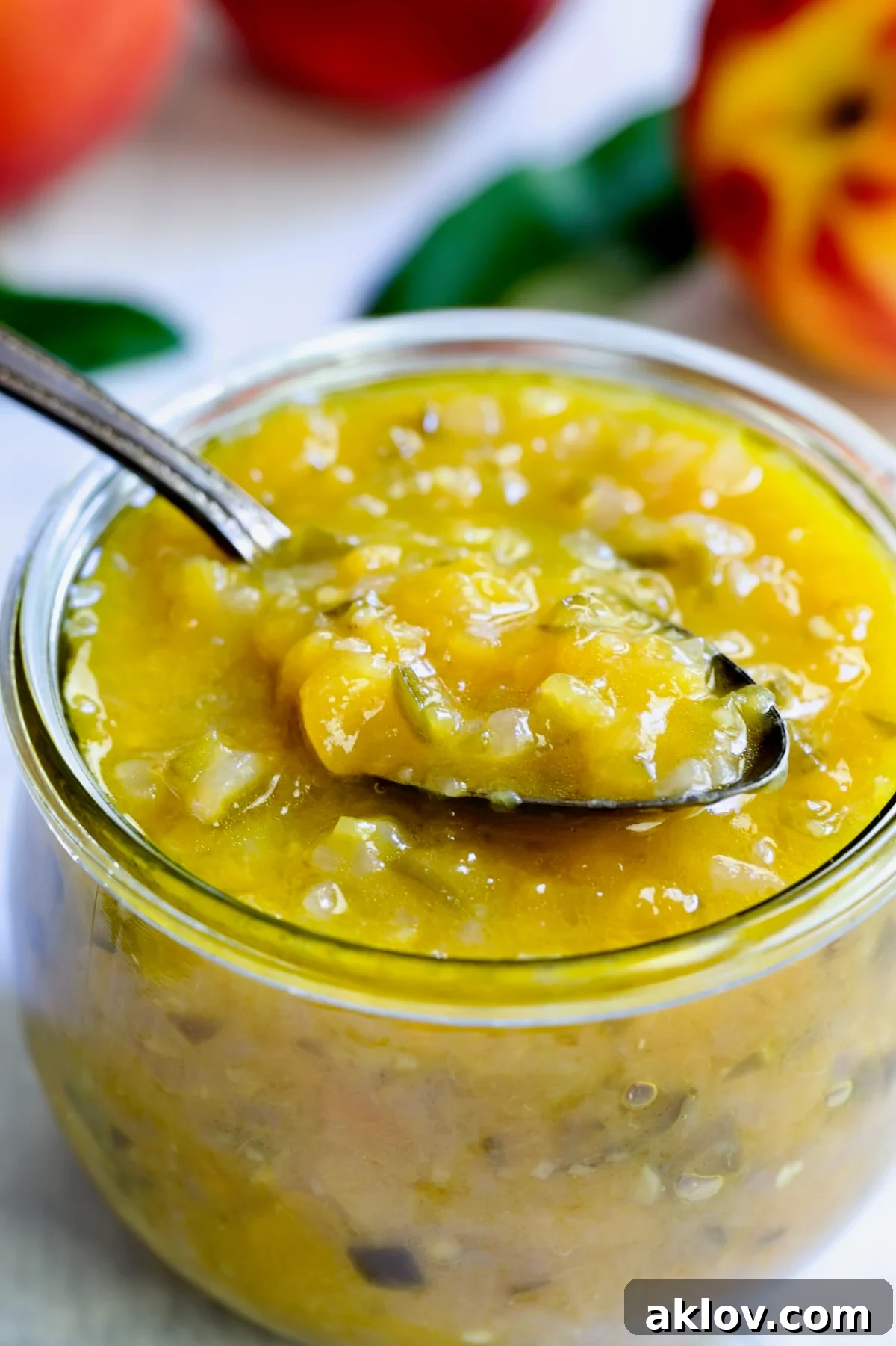 A glass jar filled with chunky, orange-hued Peach Jalapeno Chutney, with a spoon resting in it, showcasing its delicious texture.