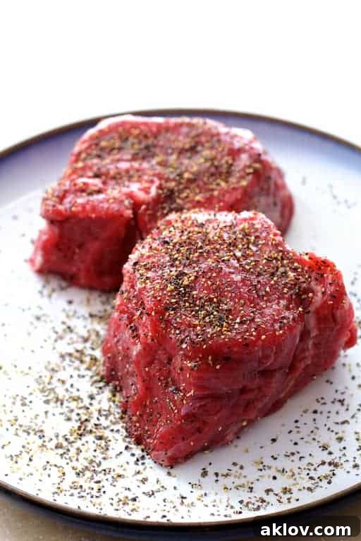 Photo of two raw uncooked beef tenderloin fillets on plate, seasoned and ready for searing.