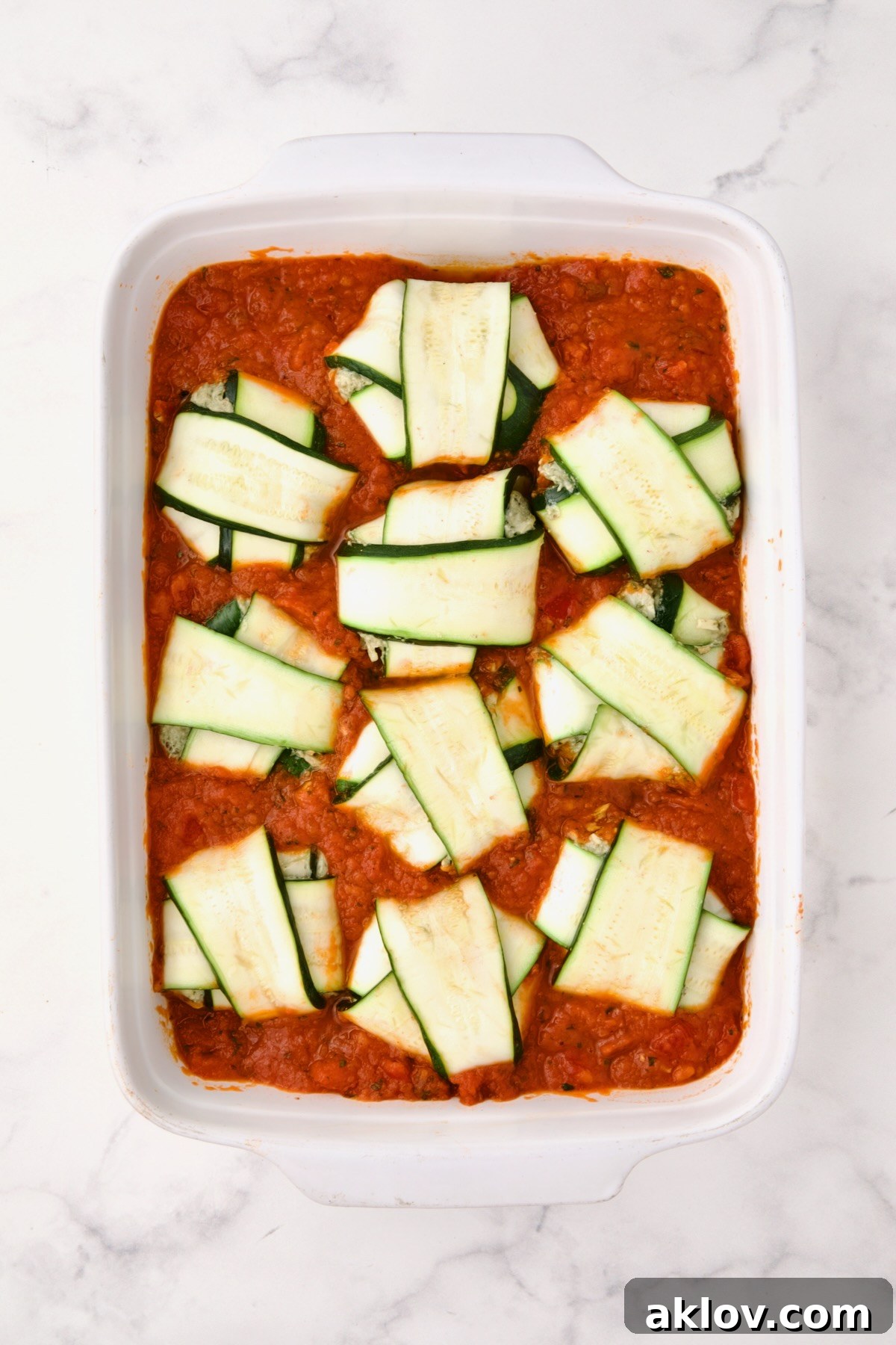 Ten assembled ravioli placed on the marinara sauce in a rectangular white ceramic baking dish before cheese is placed on the top.