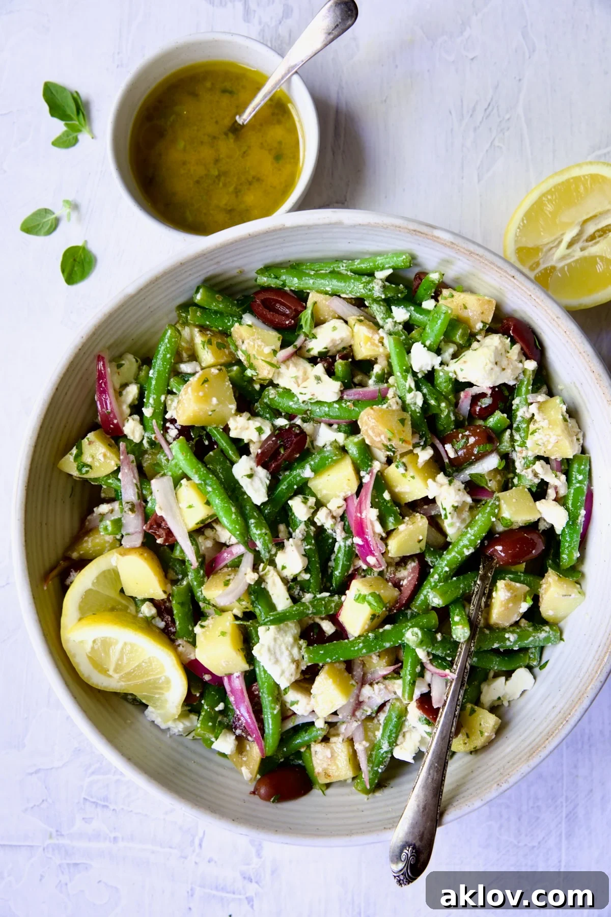 A beautifully presented Potato Green Bean Salad in a pristine white bowl, accompanied by a serving spoon, a bottle of its vibrant dressing, and a freshly cut lemon half.