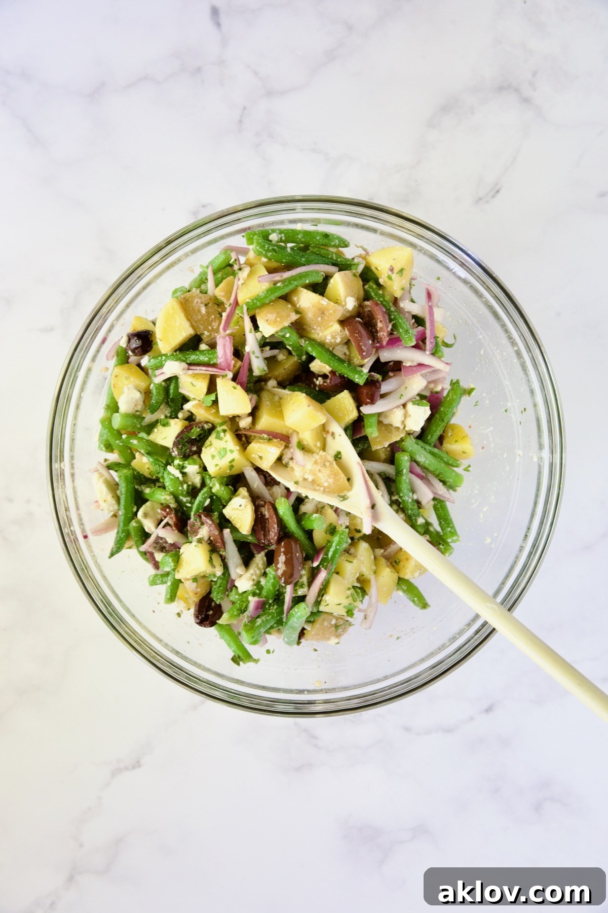 The medley of ingredients for the Potato Green Bean Salad being gently stirred together in a clear glass bowl with a white mixing spoon, ensuring even distribution of flavors.