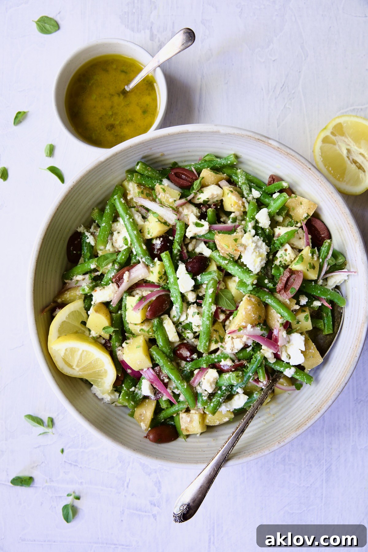 A beautifully finished Potato Green Bean Salad in a white bowl, ready for serving, with a spoon nestled within, a bottle of dressing, and a decorative cut lemon beside it.