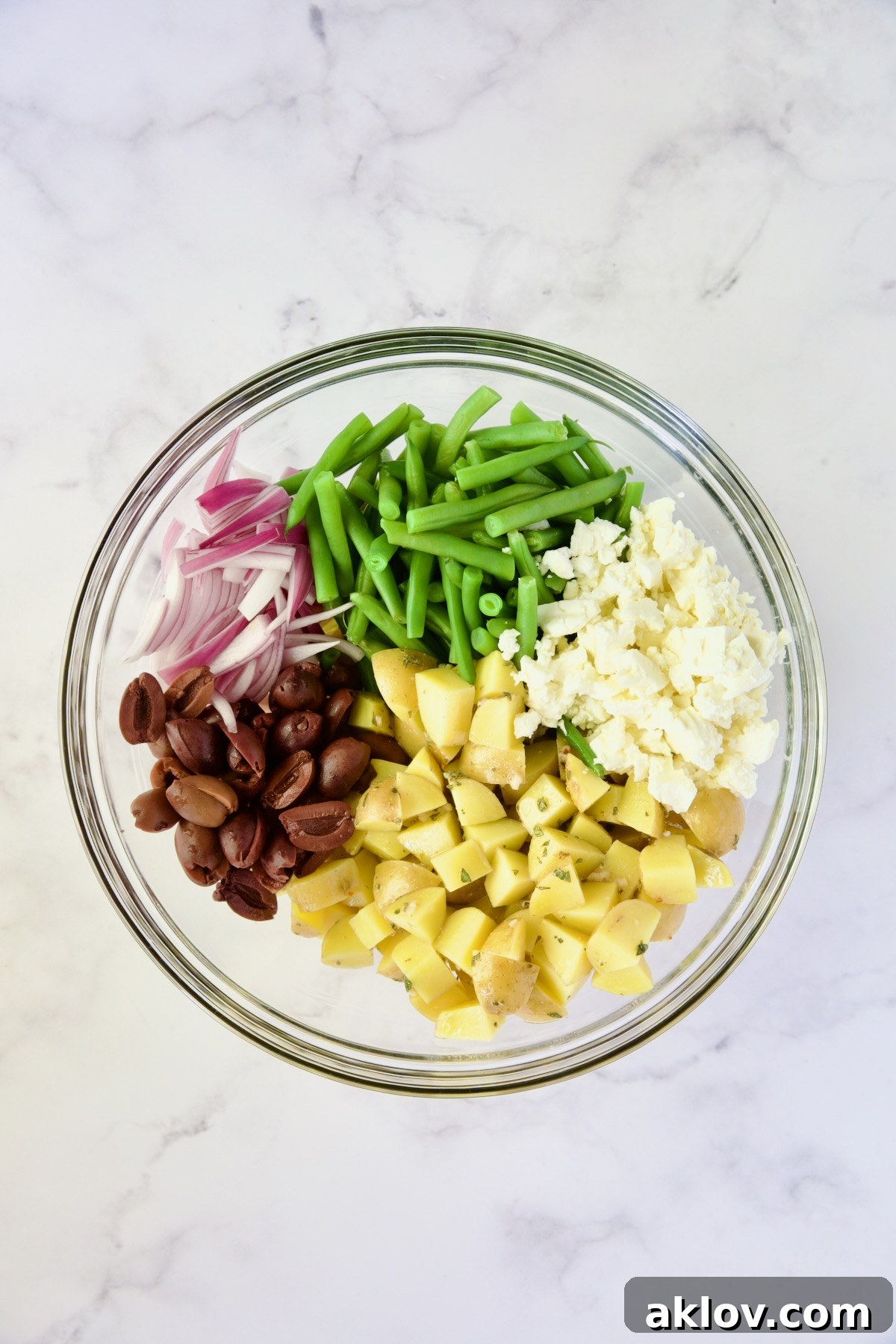 A vibrant array of fresh ingredients—red onion, blanched green beans, creamy feta cheese, perfectly cooked Yukon Gold potatoes, and briny black olives—waiting to be combined in a large glass bowl.