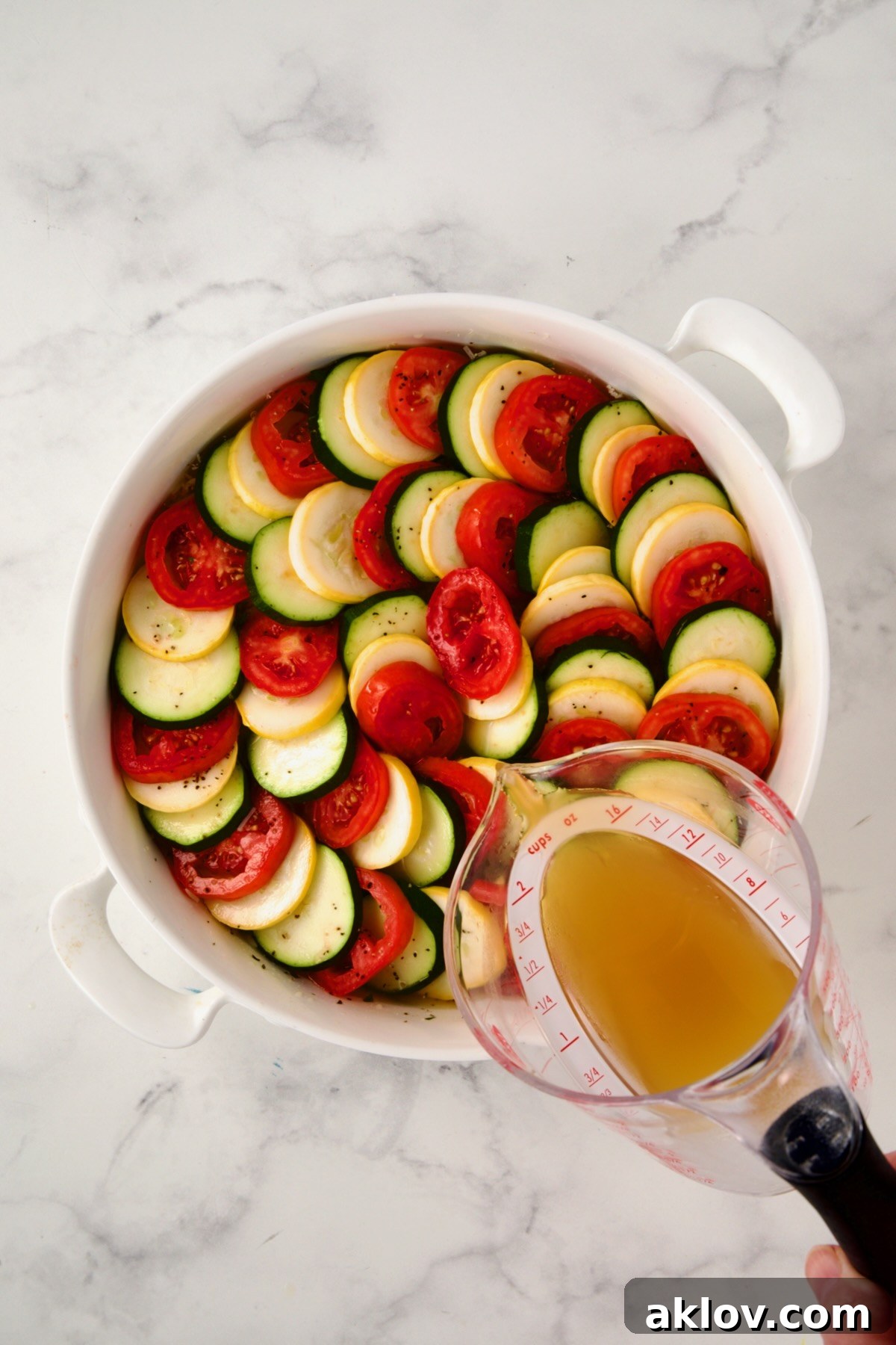 Chicken broth being gently poured over the arranged vegetables and orzo in the baking dish.