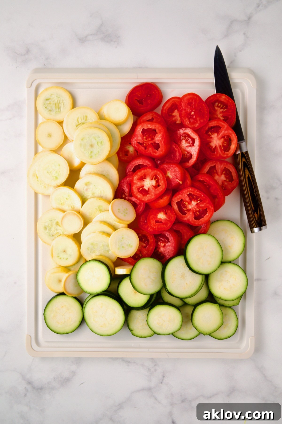 Thinly sliced yellow squash, zucchini, and tomatoes on a white cutting board, ready for seasoning.