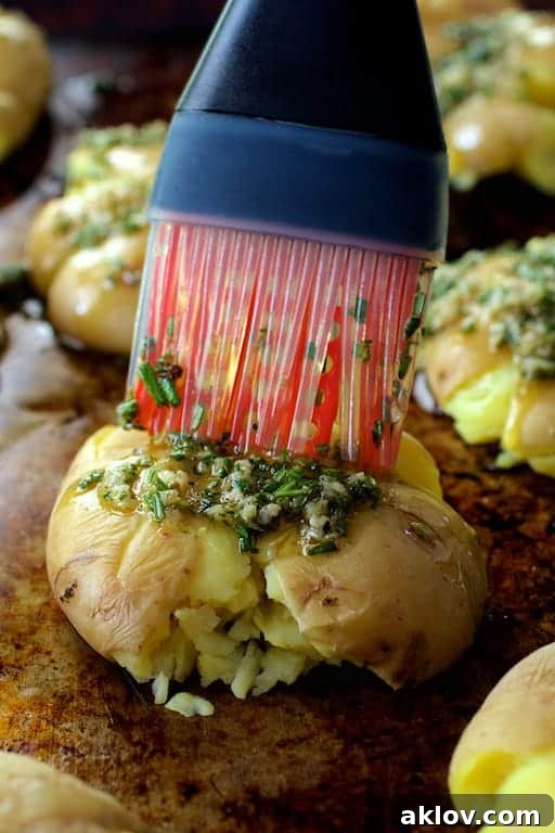 A hand using a silicone brush to generously coat crushed potatoes on a baking sheet with fragrant rosemary and garlic-infused olive oil.