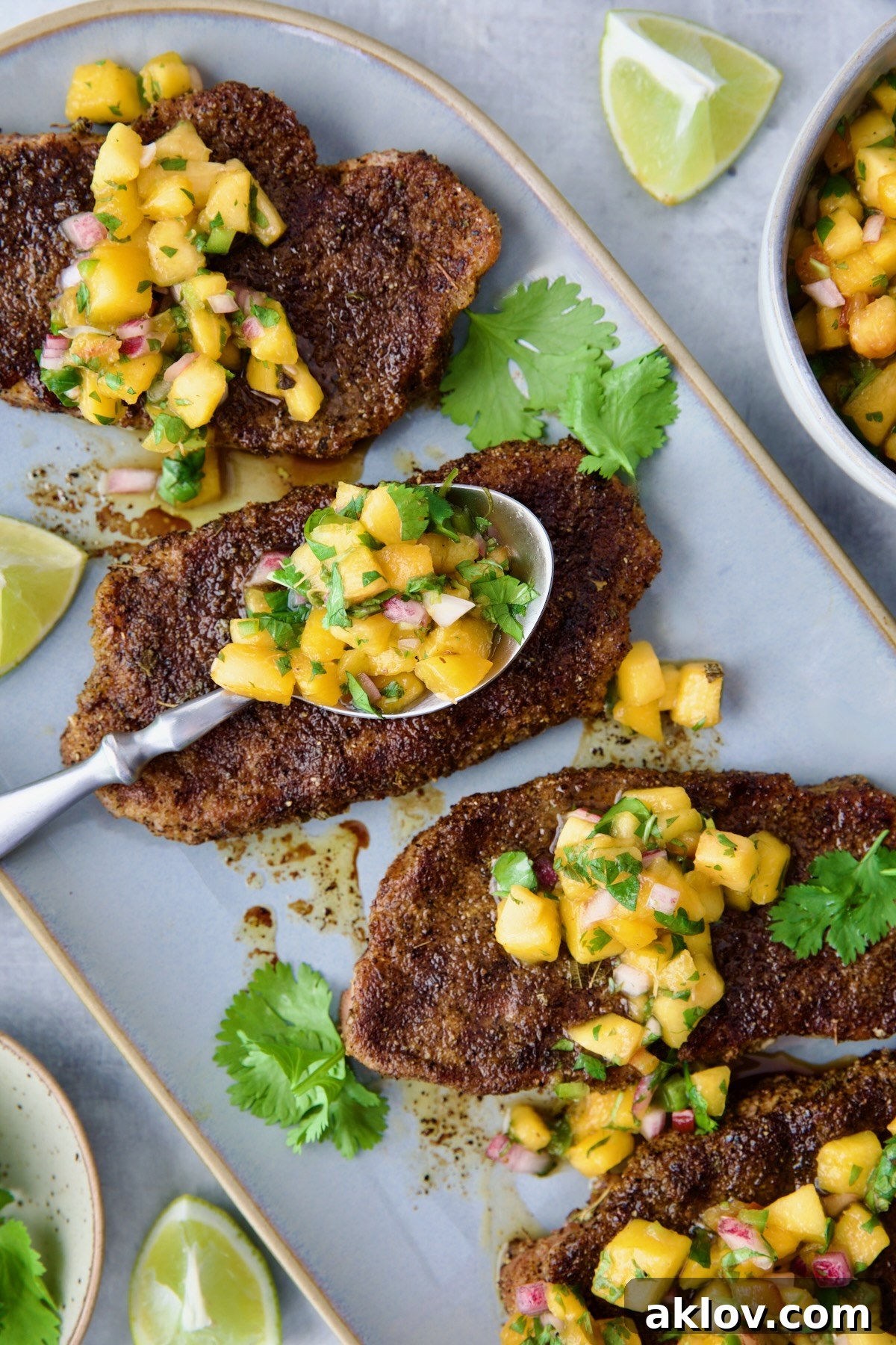 A captivating overhead shot of four grilled pork chops, each adorned with a generous serving of vibrant peach salsa, resting on an oval gray platter, with a spoon indicating a fresh dollop.
