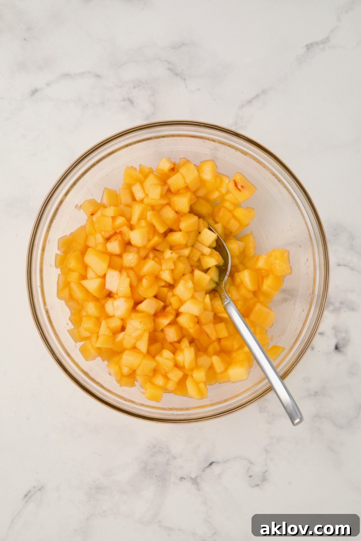 Diced peaches in glass bowl being stirred with stainless steel spoon.