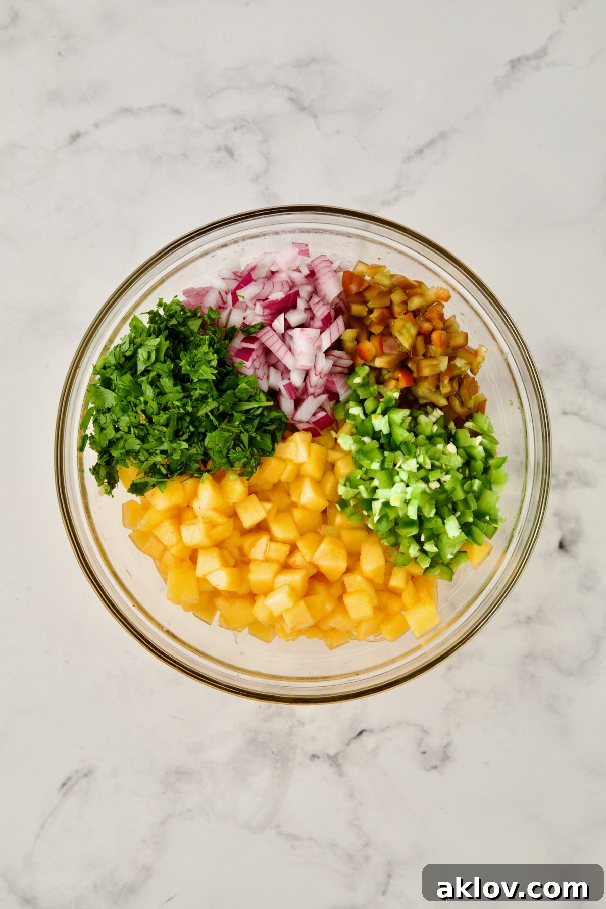 A glass bowl filled with the colorful, diced ingredients for peach salsa, including peaches, red onion, jalapeño, red bell pepper, and cilantro, before being stirred together.