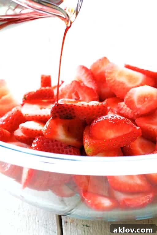 Photo of rich balsamic vinegar being gracefully poured over a bowl of vibrant red, sliced strawberries, illustrating the beginning of the maceration process.