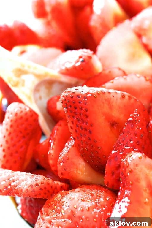 Close-up photo of bright red, sliced strawberries being gently stirred with a white spoon in a glass bowl, showcasing the process of combining ingredients for maceration.