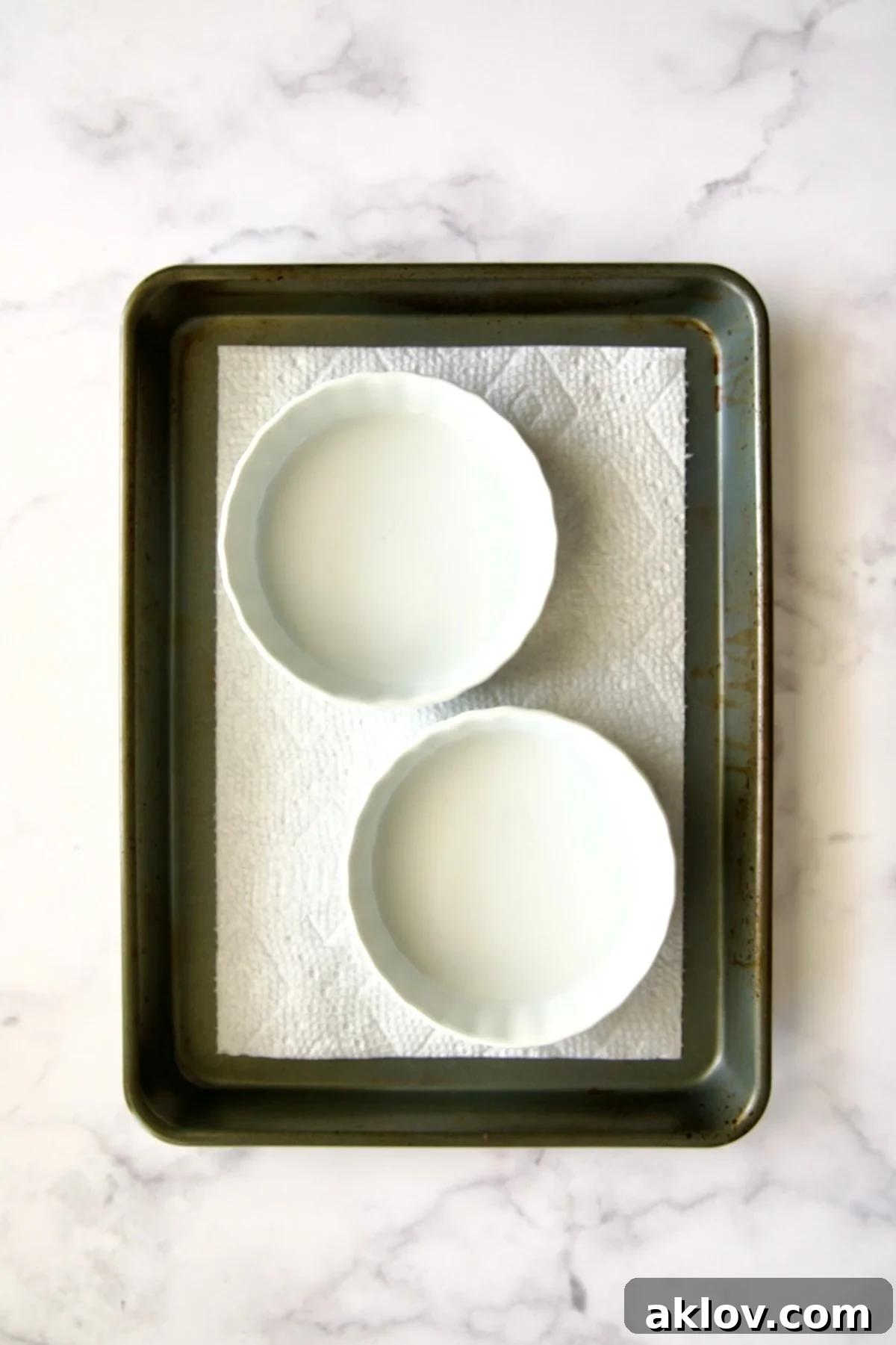 Two empty white crème brûlée dishes carefully positioned on a dry paper towel within a rimmed sheet pan, awaiting filling.
