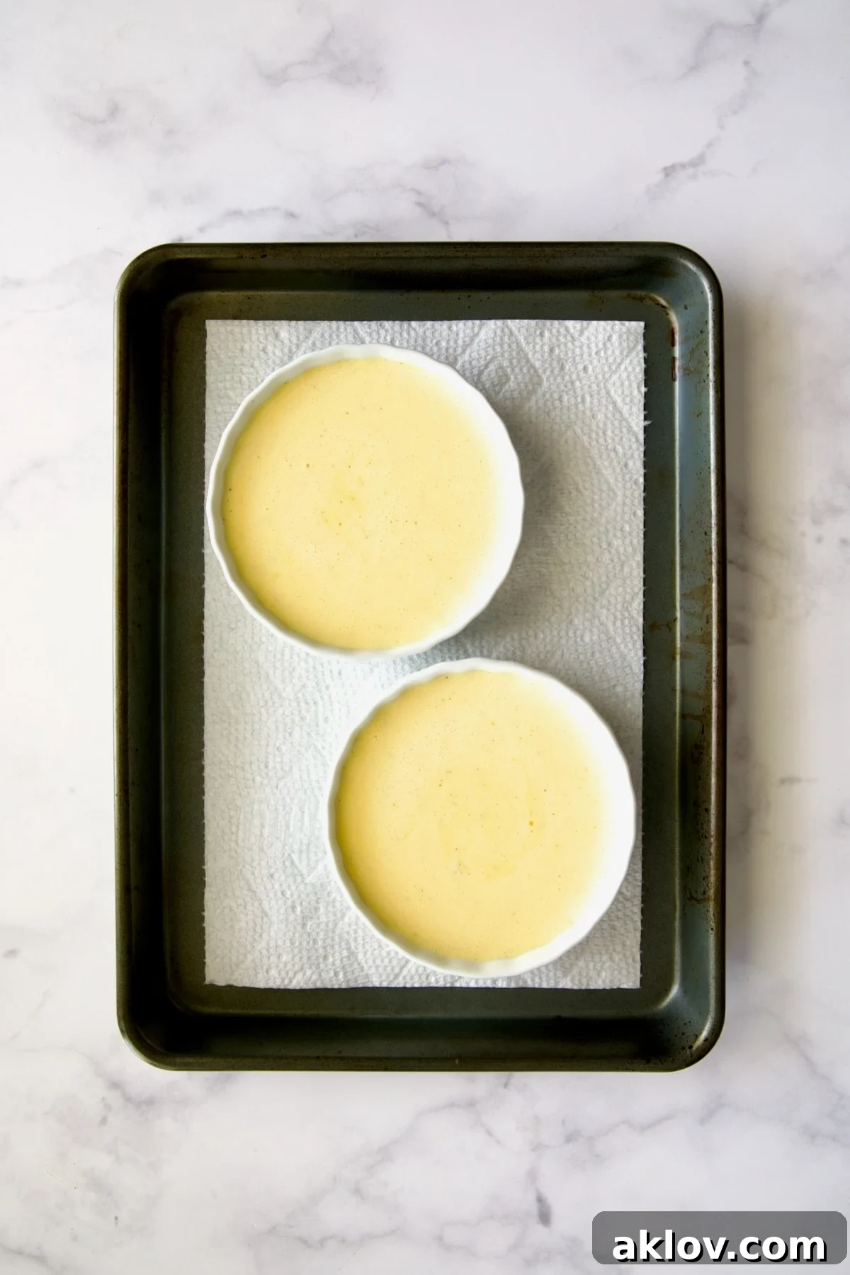 Crème brûlée batter perfectly poured into white ramekins, nestled on a paper towel in a rimmed sheet pan.