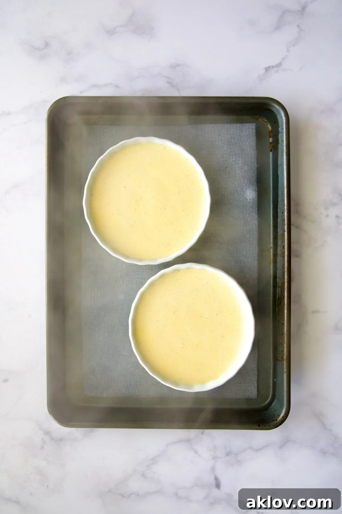 Two crème brûlée dishes in a rimmed sheet pan, with boiling water carefully added for the water bath, pre-baking.