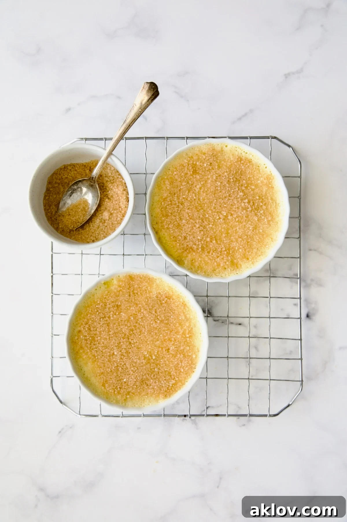 Two crème brûlées on a wire rack, with turbinado sugar evenly sprinkled over their tops, alongside a bowl of extra sugar.