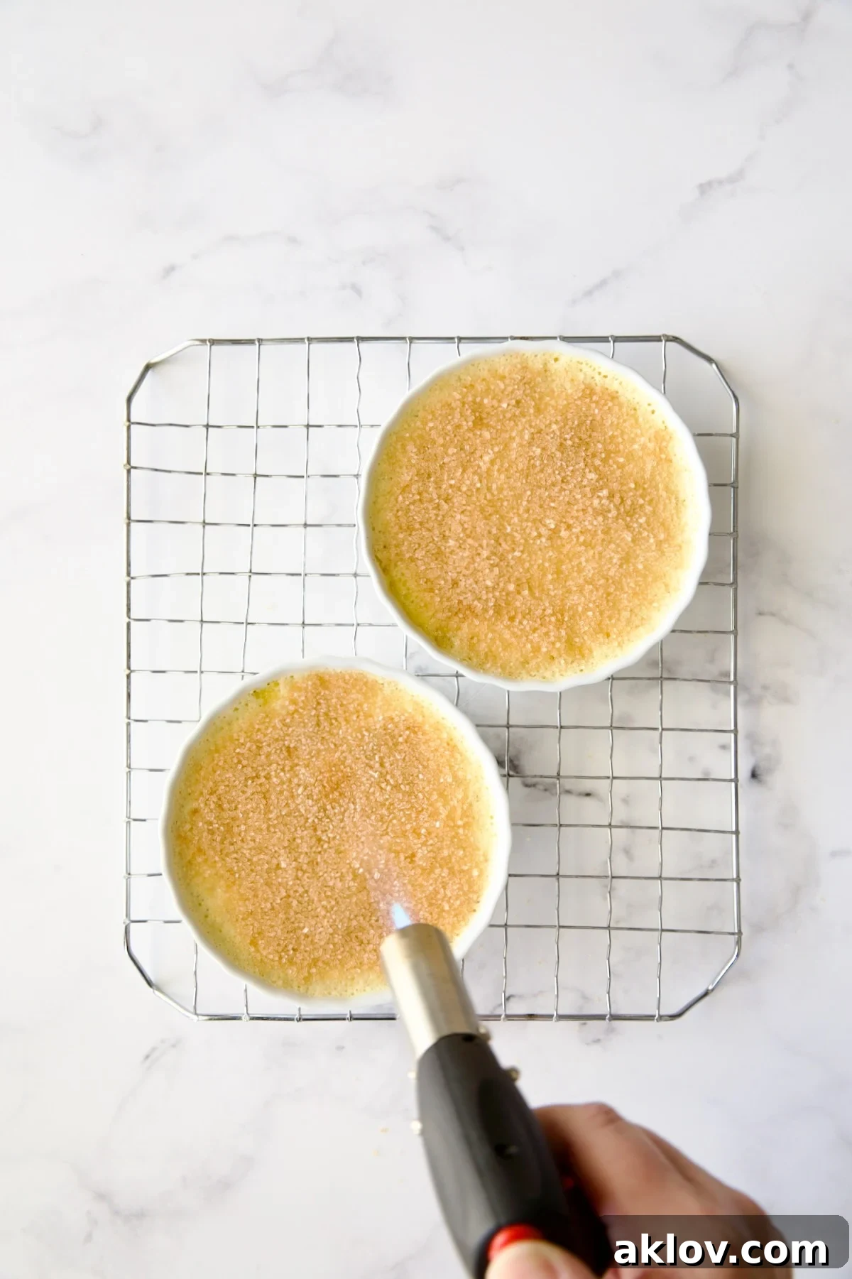 A culinary torch poised to caramelize the turbinado sugar topping on two crème brûlées set on a wire rack, creating the signature crust.