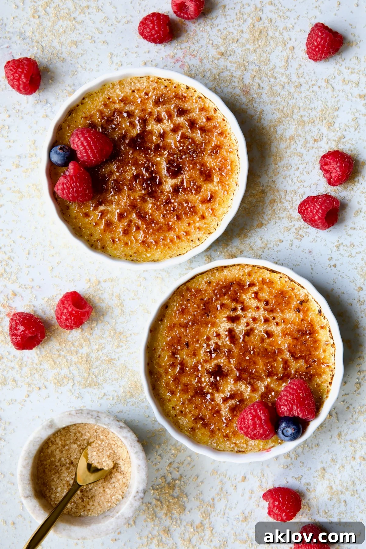 Two dishes of Classic Crème Brûlée for Two, elegantly garnished with red raspberries and blueberries, accompanied by a small bowl of turbinado sugar.