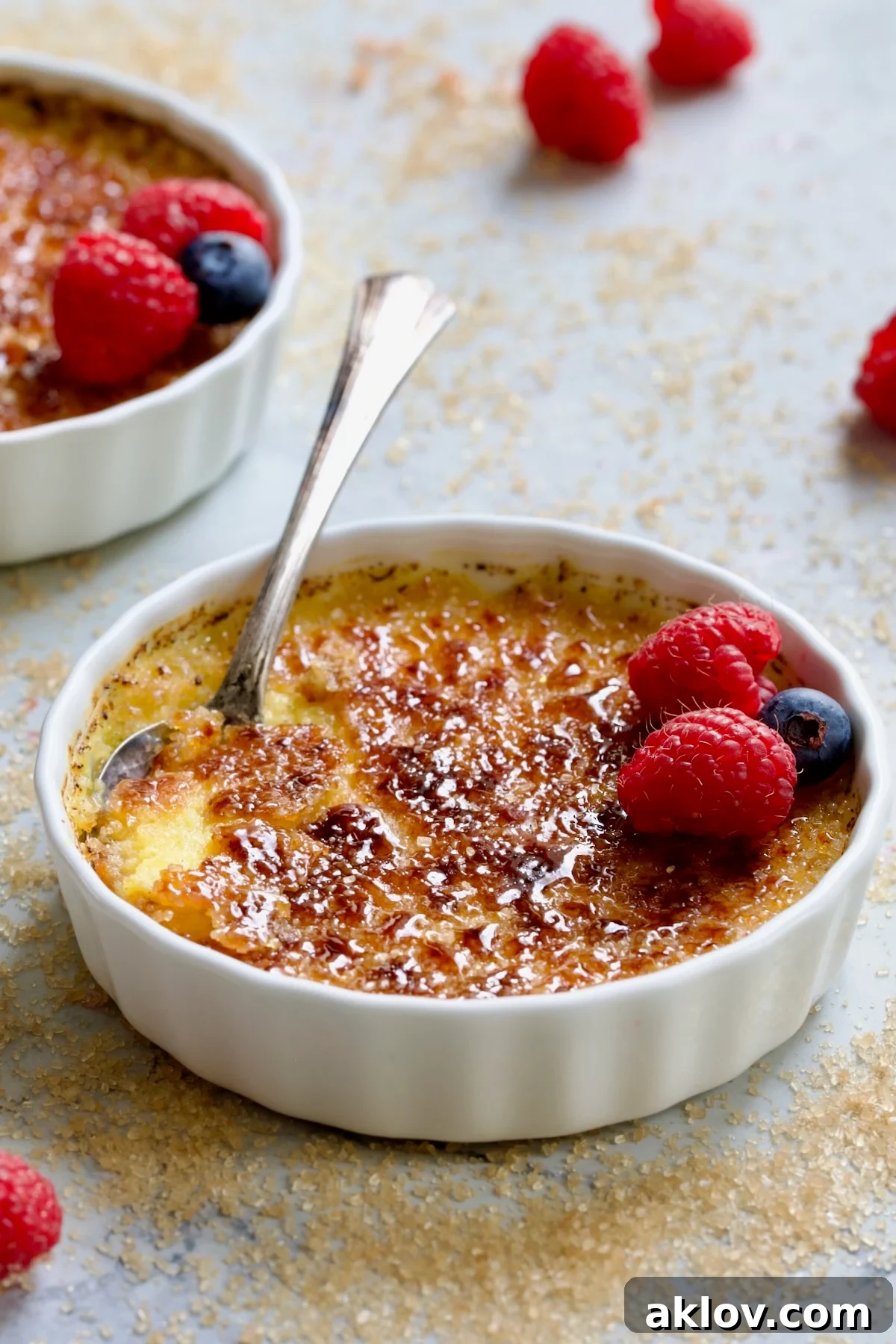 One dish of Classic Crème Brûlée for Two on a subtly sugary surface, garnished with fresh red raspberries and blueberries, highlighting its inviting texture.