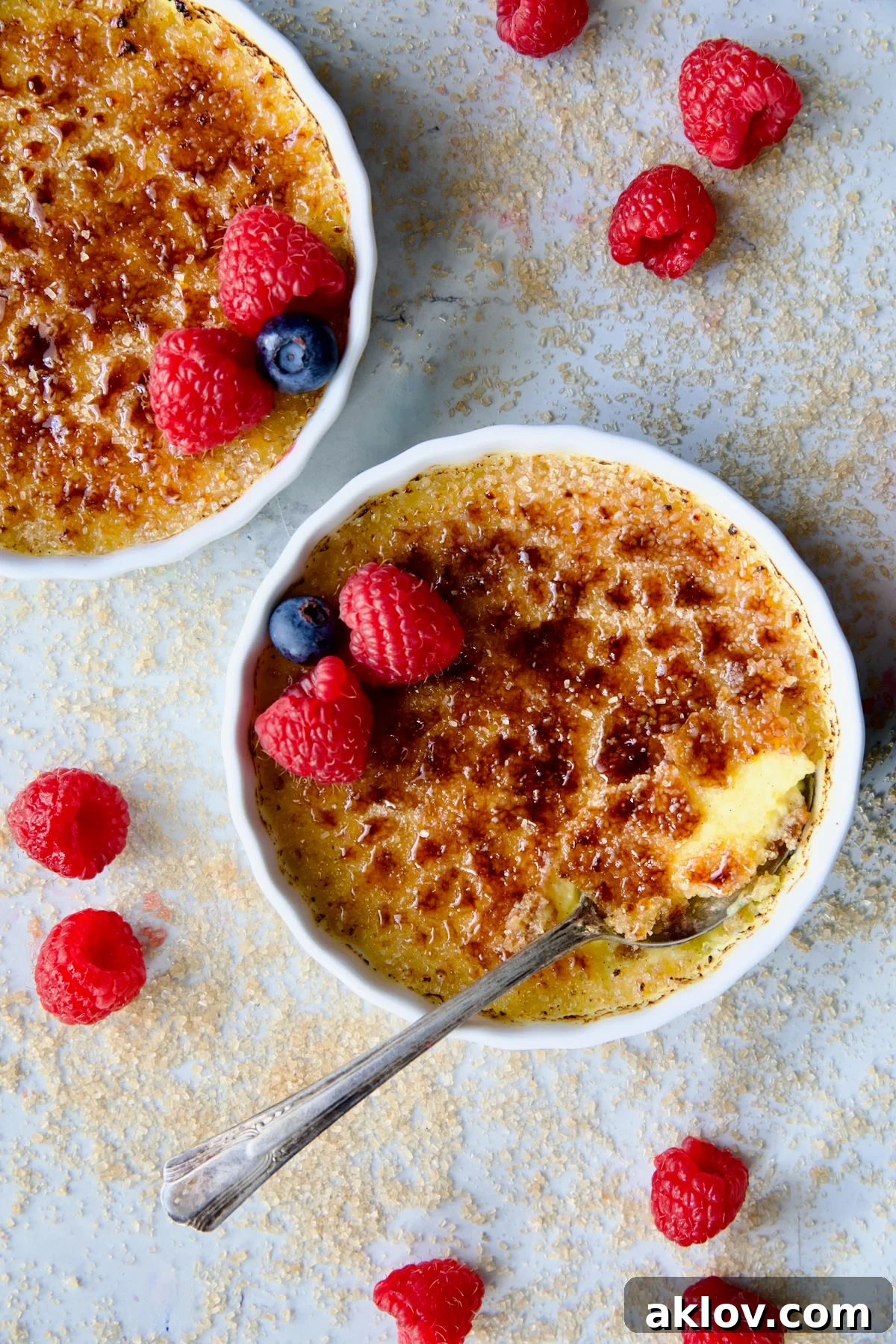 Two dishes of Classic Crème Brûlée for Two, adorned with red raspberries and blueberries, with a spoon gracefully breaking into one of the caramelized crusts, revealing the creamy custard beneath.