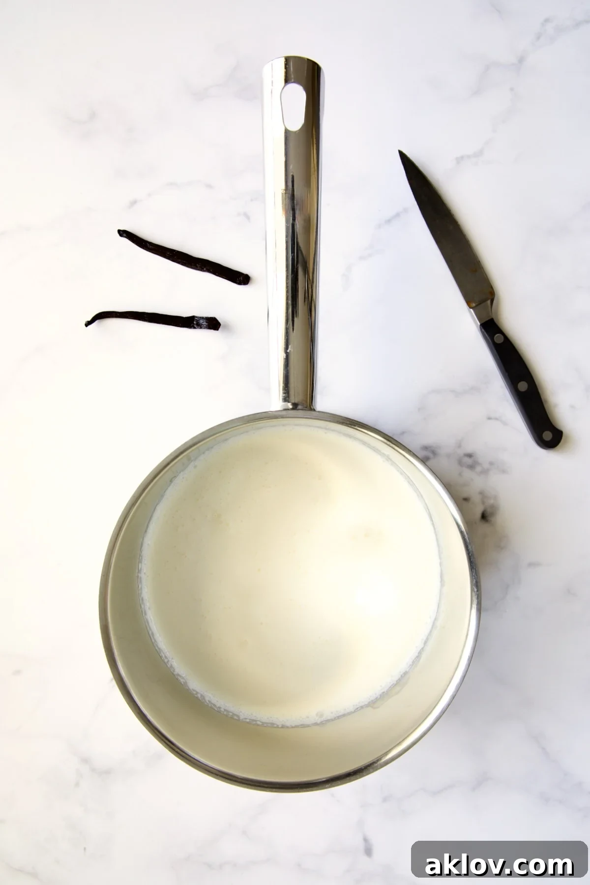 Heavy cream warming in a stainless steel saucepan, with a halved vanilla bean and paring knife nearby for preparation.