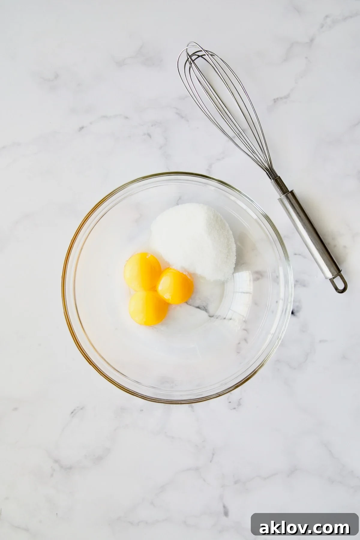 Three golden egg yolks and granulated sugar waiting to be combined in a clear glass bowl, with a wire whisk poised for action.