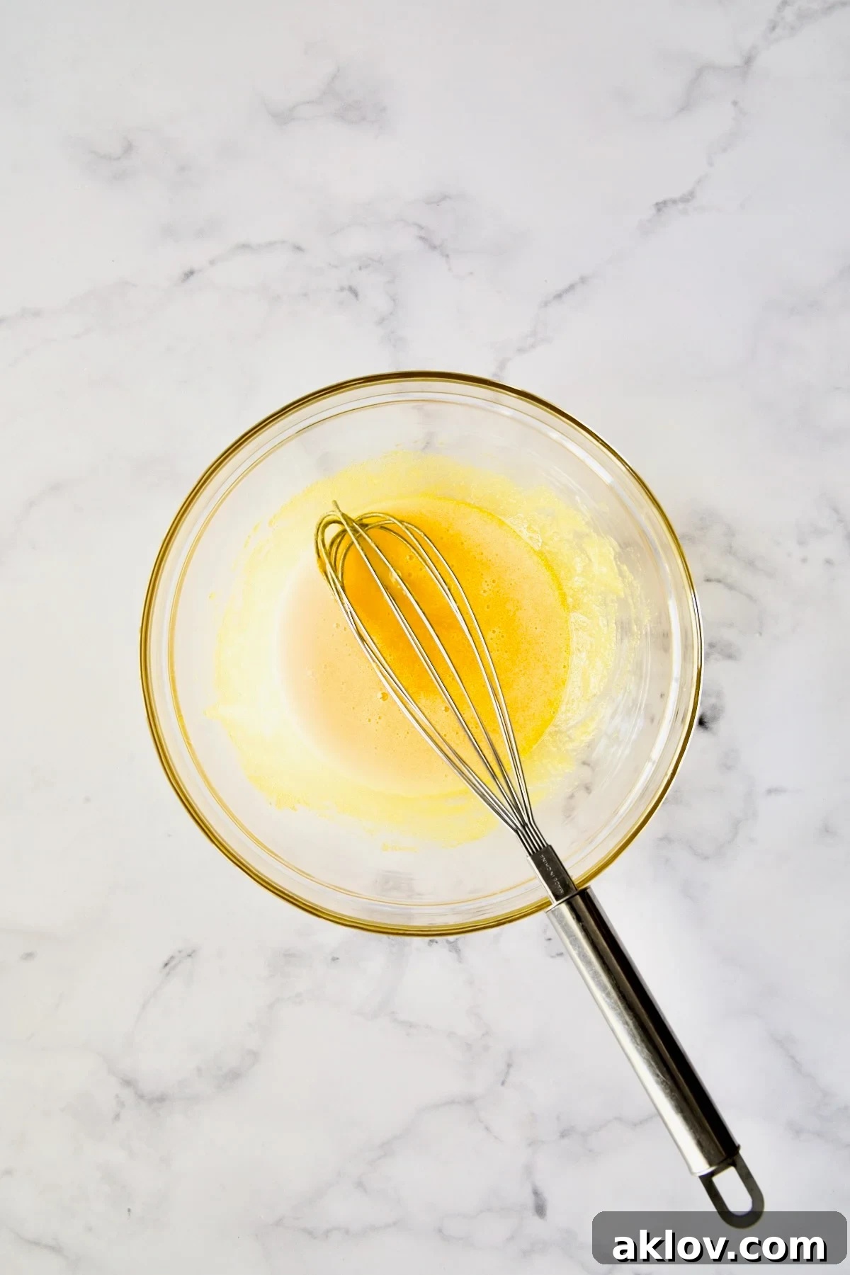 A wire whisk actively beating egg yolks and sugar together in a glass bowl, illustrating the initial mixing process.