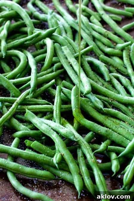 Close-up process photo of fresh green beans spread evenly on a baking sheet, being drizzled with high-quality olive oil.