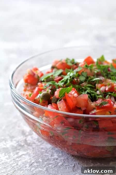A vibrant Tomato Caper Relish, showcasing finely diced tomatoes, capers, and herbs, expertly mixed in a clear glass bowl on a clean white surface.