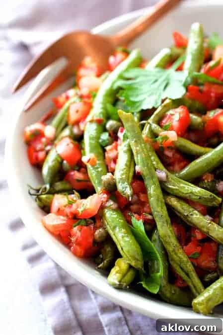 A close-up shot of the finished Roasted Green Beans with Tomato Caper Relish in a white serving bowl, ready to be enjoyed with a copper serving fork.