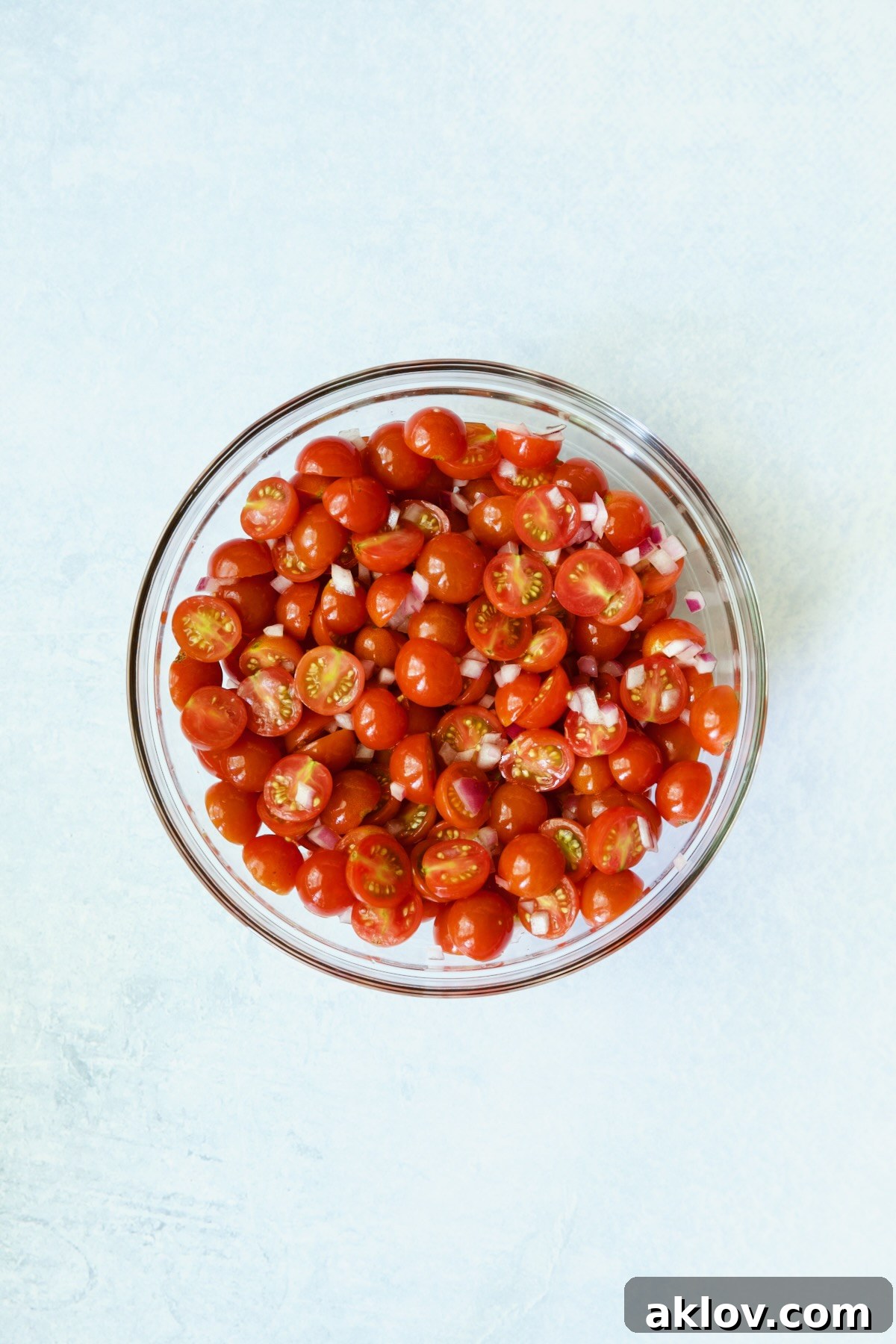 Halved cherry tomatoes and diced red onion in a clear glass bowl, seasoned and ready for maceration.