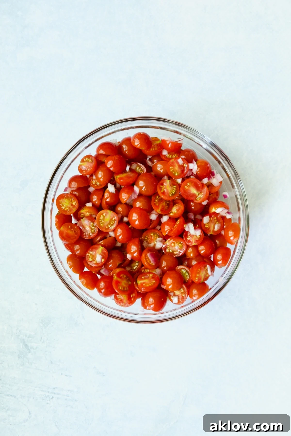 A close-up shot of halved cherry tomatoes and diced red onion resting in a clear glass bowl, seasoned and awaiting the next step.