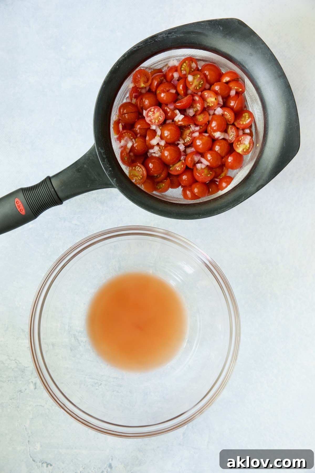 Cherry tomatoes and red onion draining in a sieve, with their flavorful juices collected in a glass bowl below.