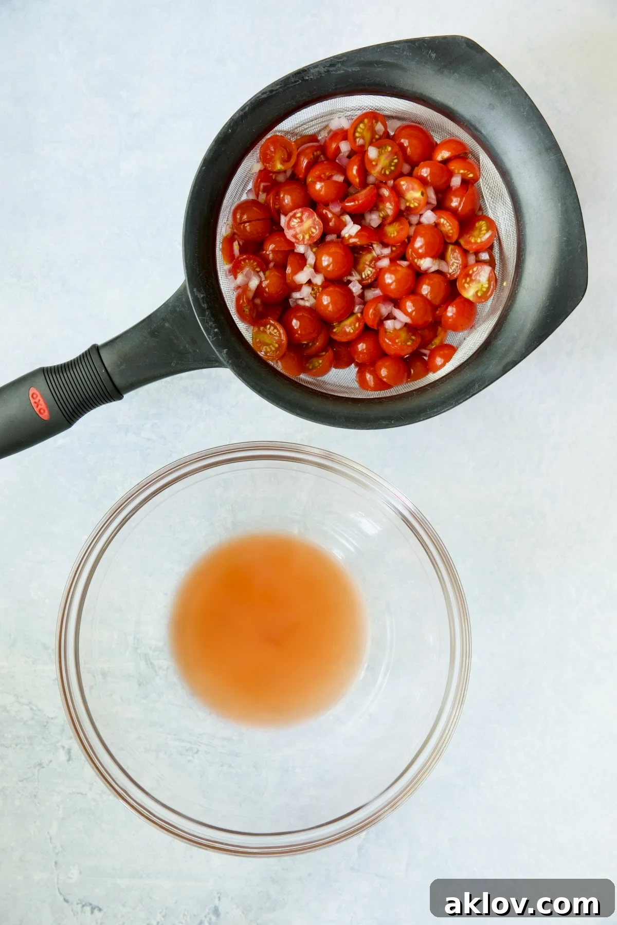 A sieve holding marinated cherry tomatoes and red onion, positioned over a glass bowl to collect the reserved flavorful juices.