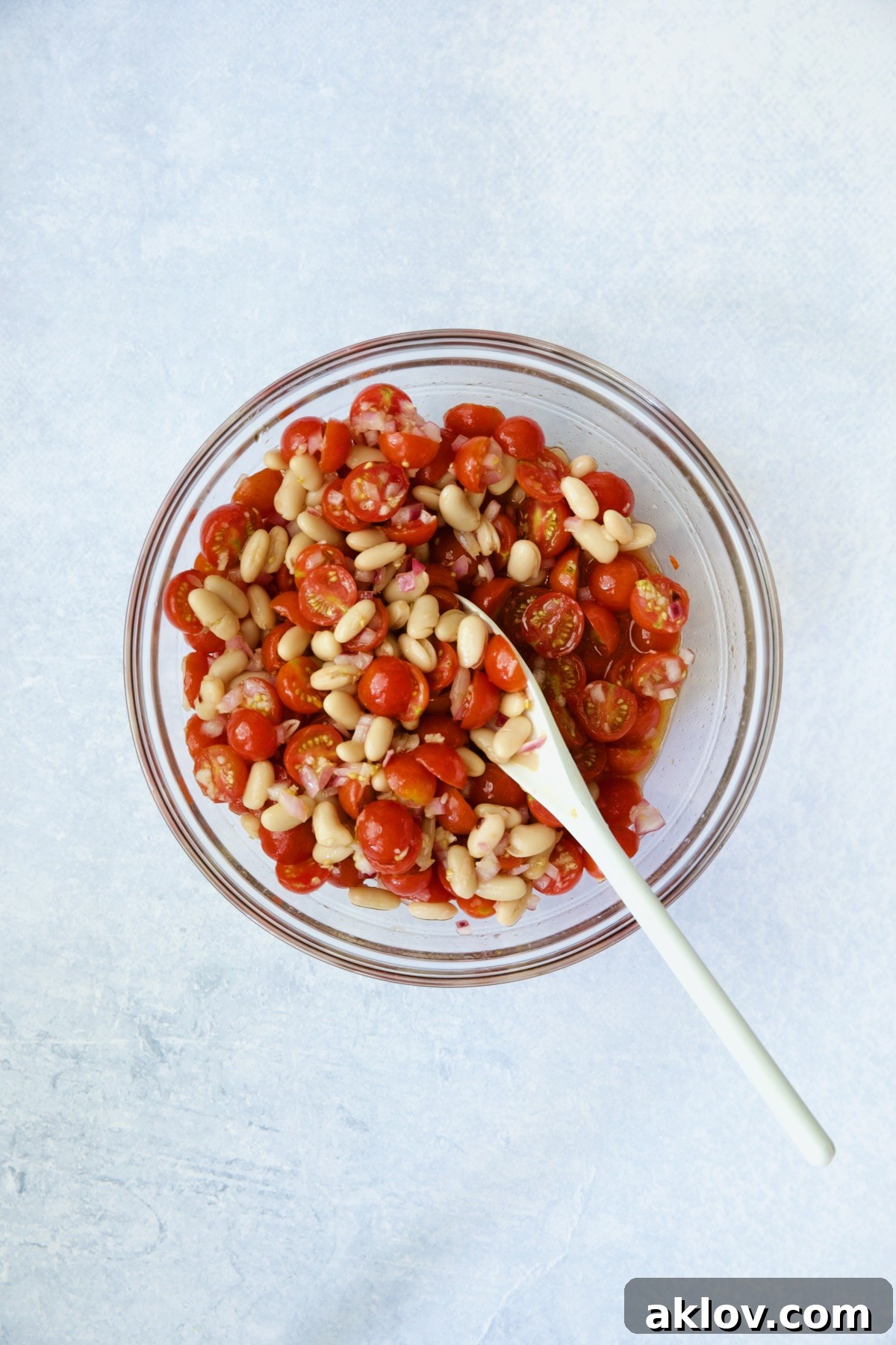 A glass bowl containing white beans and the dressing being stirred with a white spoon.