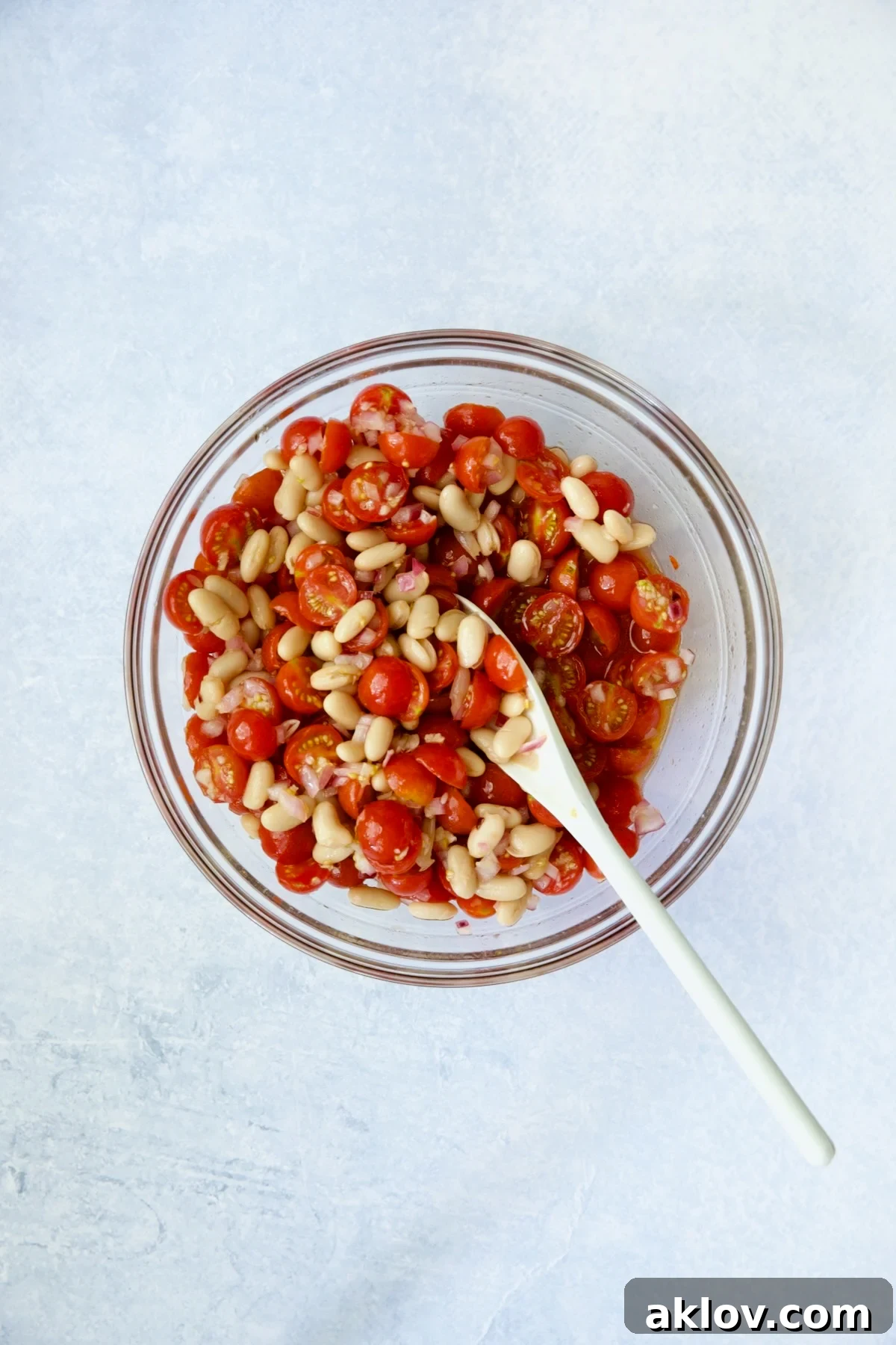 A white spoon gently stirring the salad ingredients – tomatoes, onions, beans, and dressing – in a large glass bowl, ensuring even coating.