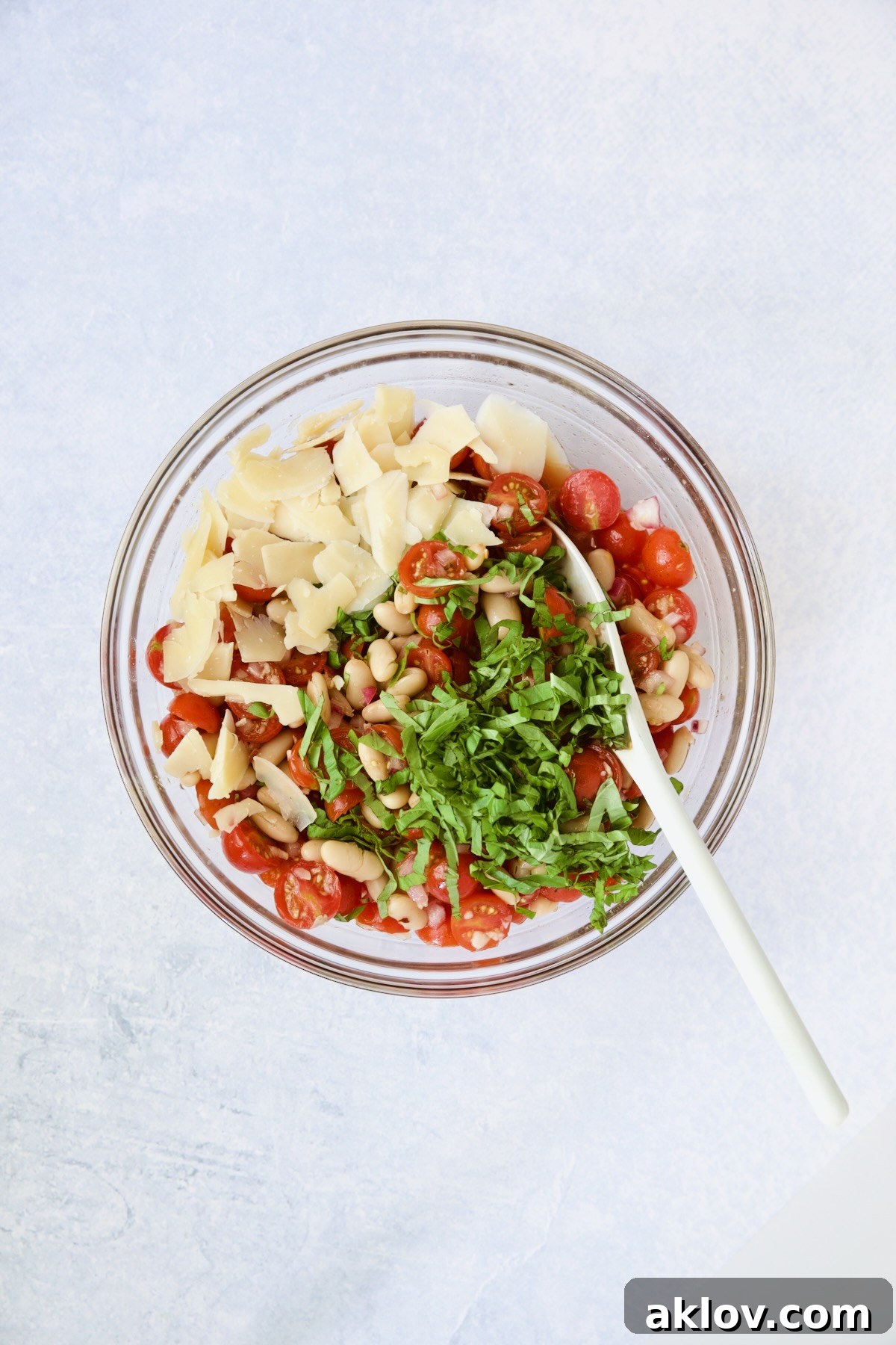 The nearly complete salad in a glass bowl, with basil and Parmesan added, being gently stirred with a white spoon.