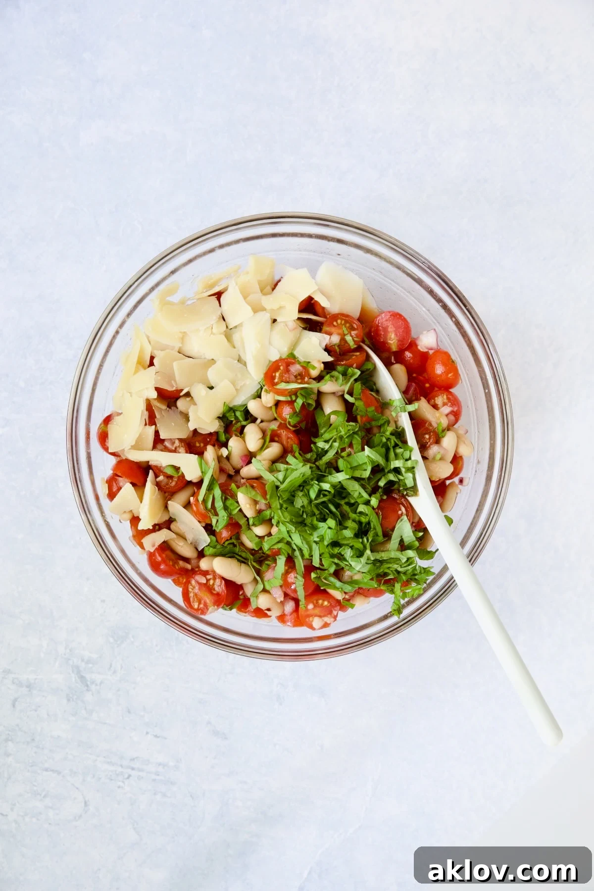 The nearly finished salad in a glass bowl, being gently mixed with a white spoon after the addition of fresh basil and Parmesan cheese.