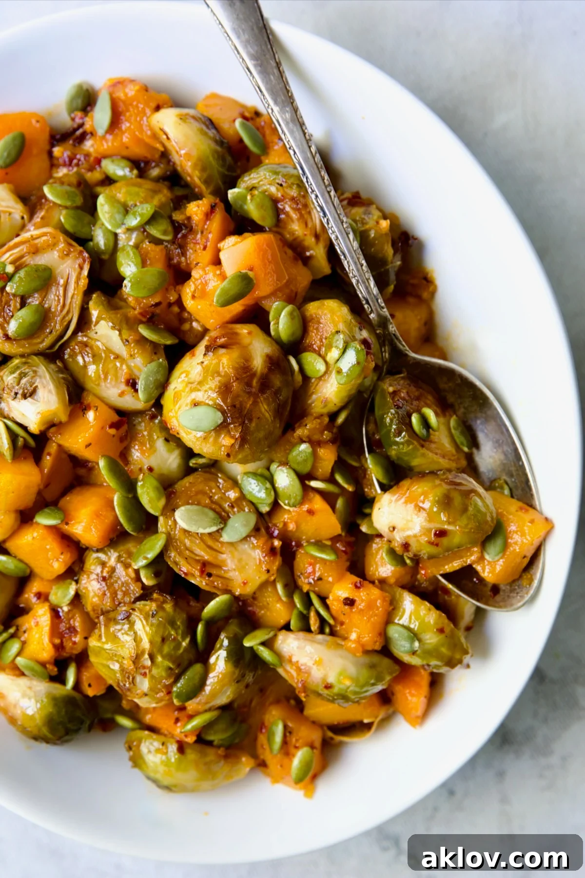 Vibrantly colored Maple Chili Crisp Brussels Sprouts and Butternut Squash, generously garnished with toasted pepitas, presented in an elegant white serving bowl with a wooden spoon.