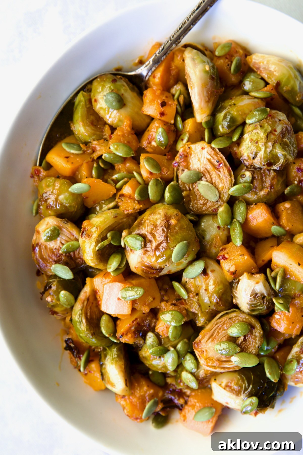 A final, mouth-watering shot of Maple Chili Crisp Brussels Sprouts and Butternut Squash, artfully presented with a sprinkle of pepitas, emphasizing its inviting texture and vibrant colors in a white serving bowl.