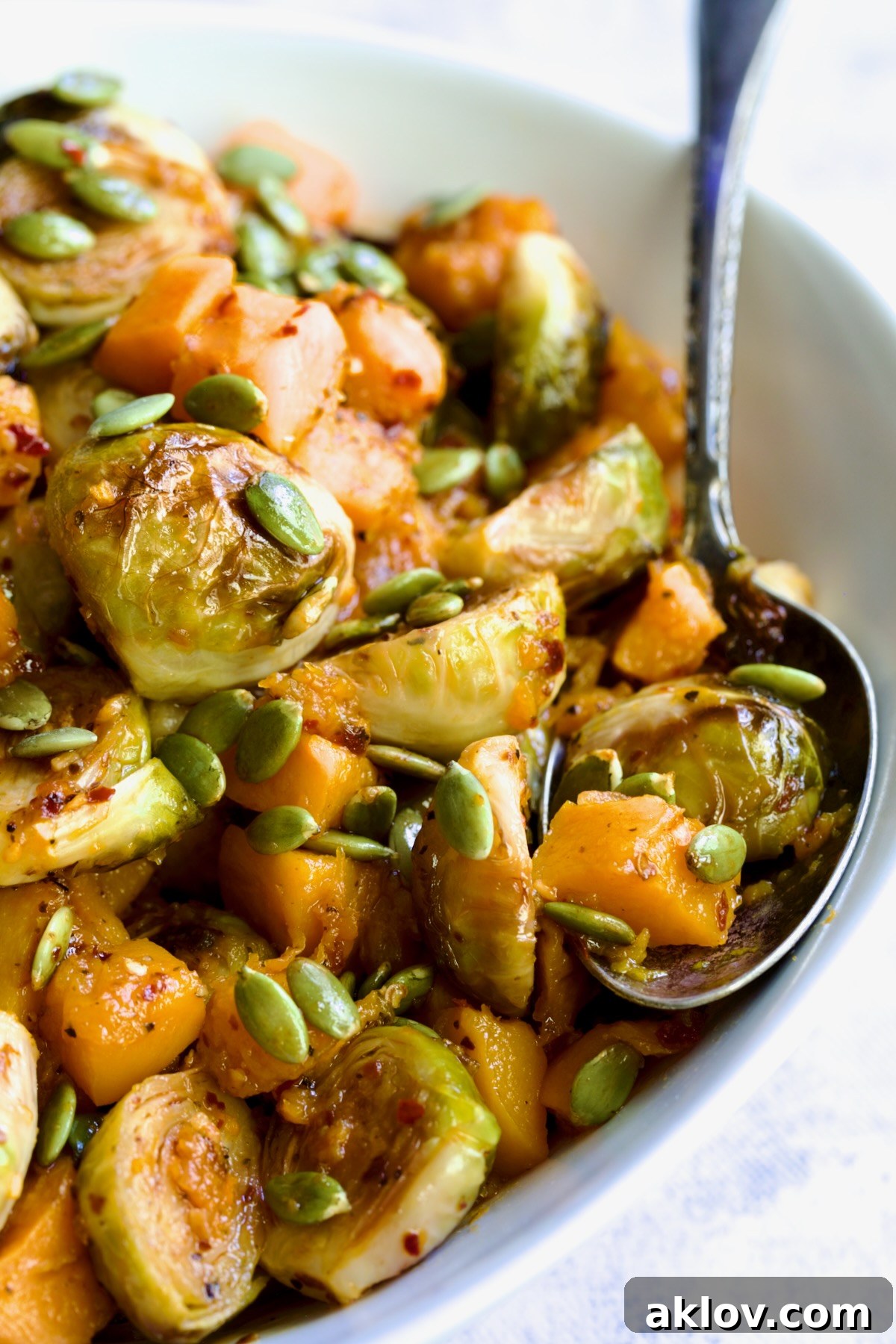 A close-up view of Maple Chili Crisp Brussels Sprouts and Butternut Squash, sprinkled with pepitas, served in a pristine white bowl alongside a wooden serving spoon, highlighting the dish's vibrant colors and textures.