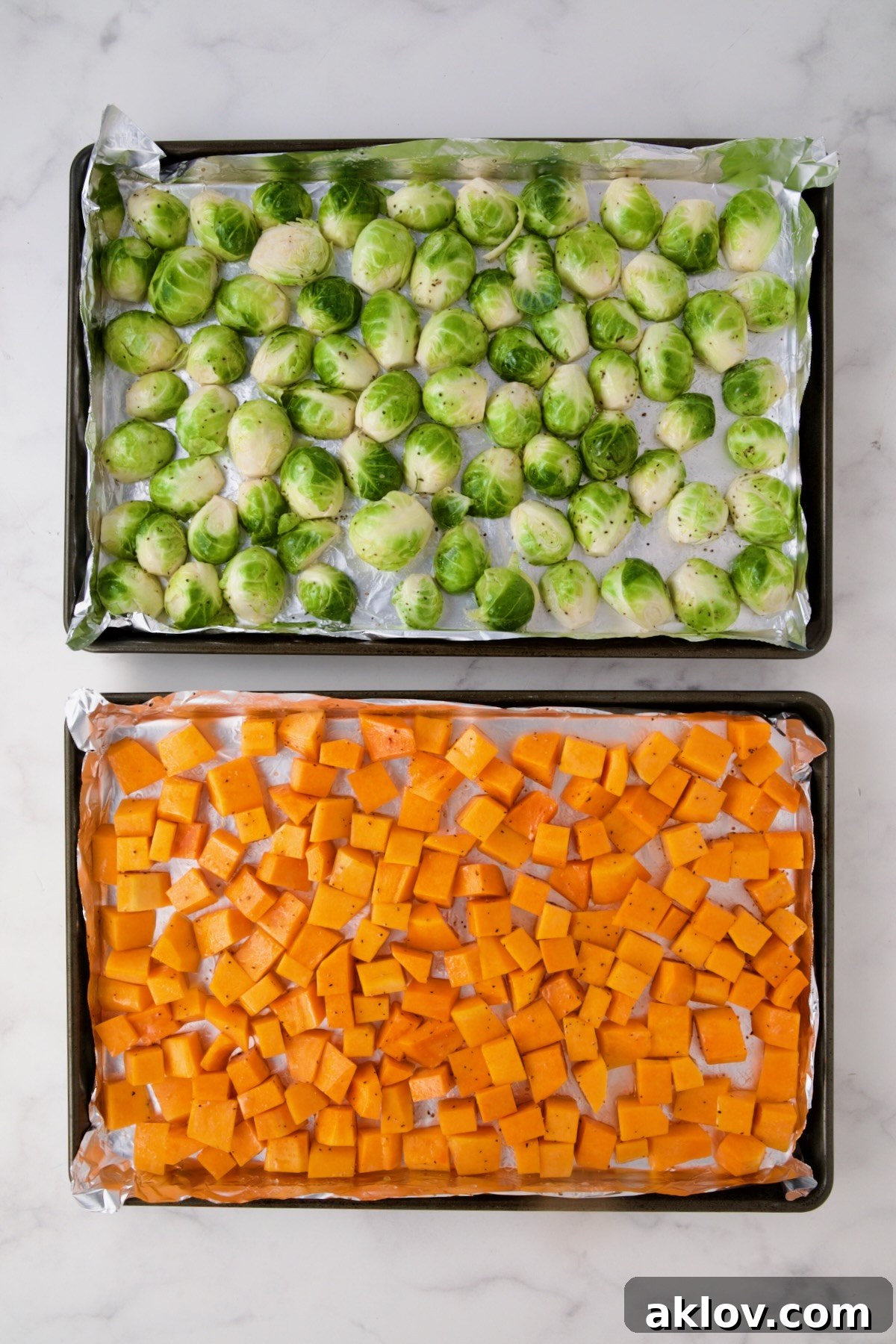 Freshly halved Brussels sprouts and uniformly cubed butternut squash are spread out on separate nonstick aluminum foil-lined rimmed baking sheets, perfectly prepped and ready for roasting.