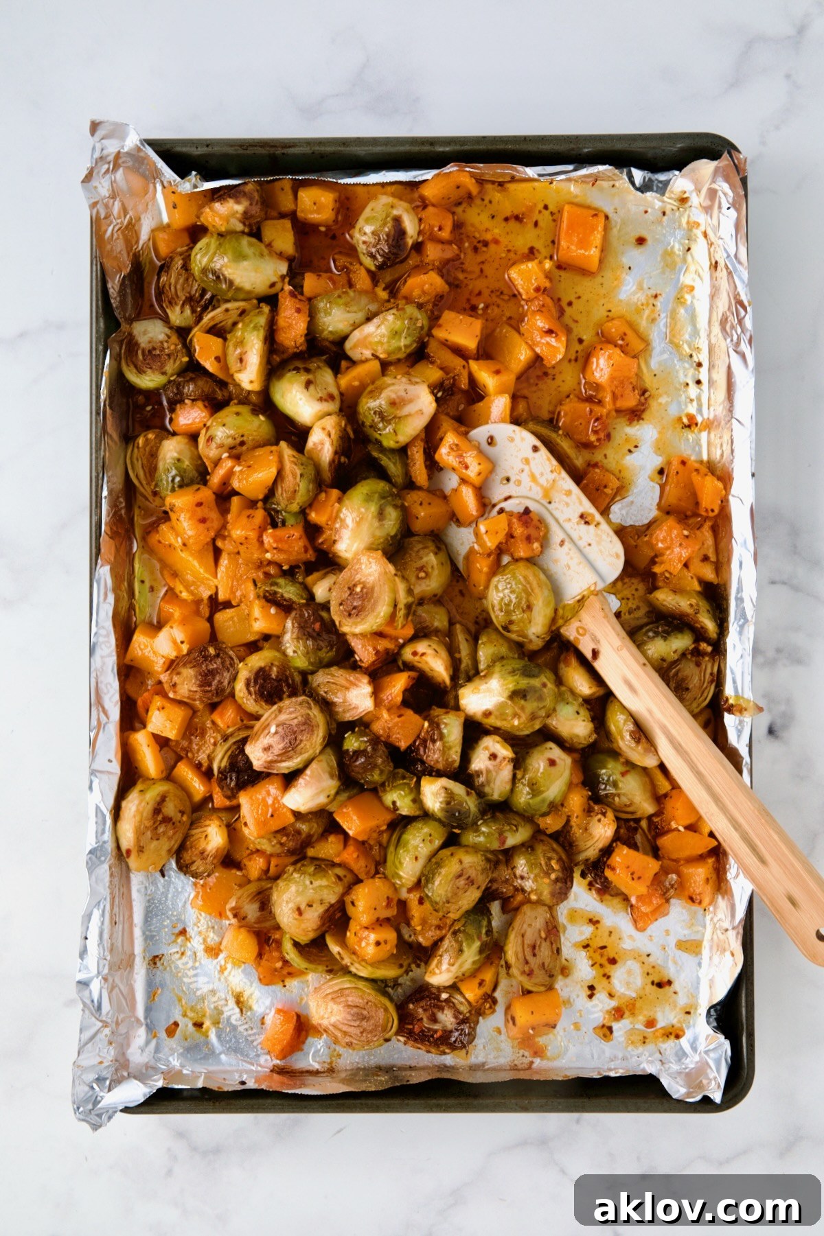The fully combined and sauce-coated roasted Brussels sprouts and butternut squash are stirred with a wooden spatula on a foil-lined baking sheet, glistening and ready for a final quick roast.