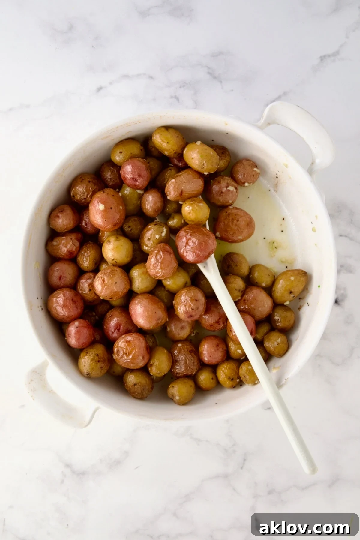 White mixing spoon stirring butter and garlic into mini potatoes in round white baking dish.