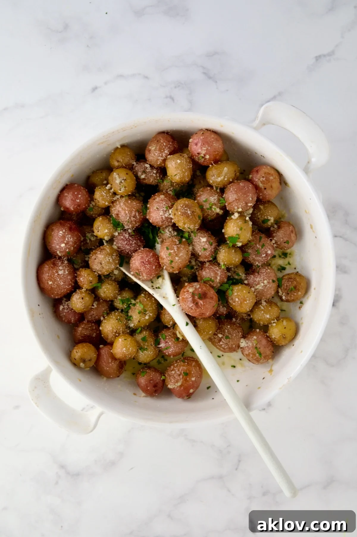 Parsley added to roasted mini potatoes and being stirred with white mixing spoon in round white baking dish.