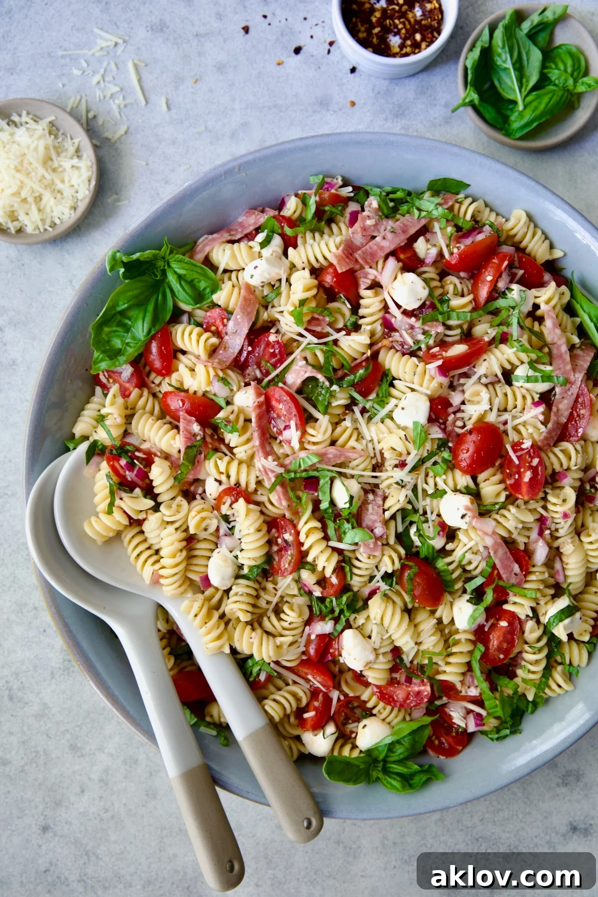 Caprese Pasta Salad in blue bowl with serving utensils with small bowls of Parmesan cheese, crushed red pepper flakes and fresh basil near it.