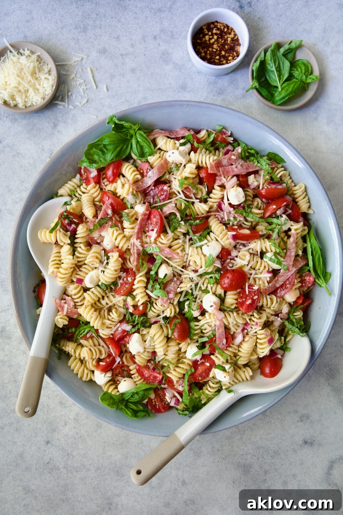 Caprese Pasta Salad in blue bowl with serving utensils with small bowls of Parmesan cheese, crushed red pepper flakes and fresh basil near it.