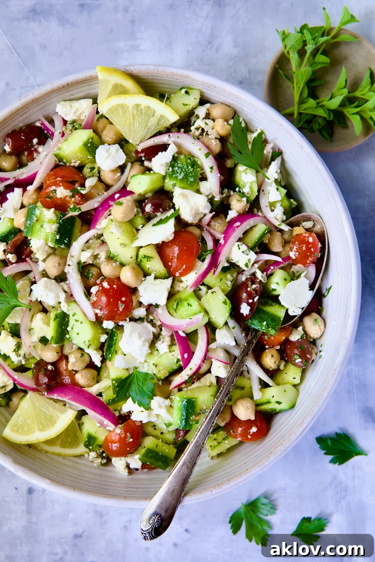 Greek Cucumber Salad in white bowl with serving spoon with oregano and parsley scattered about.