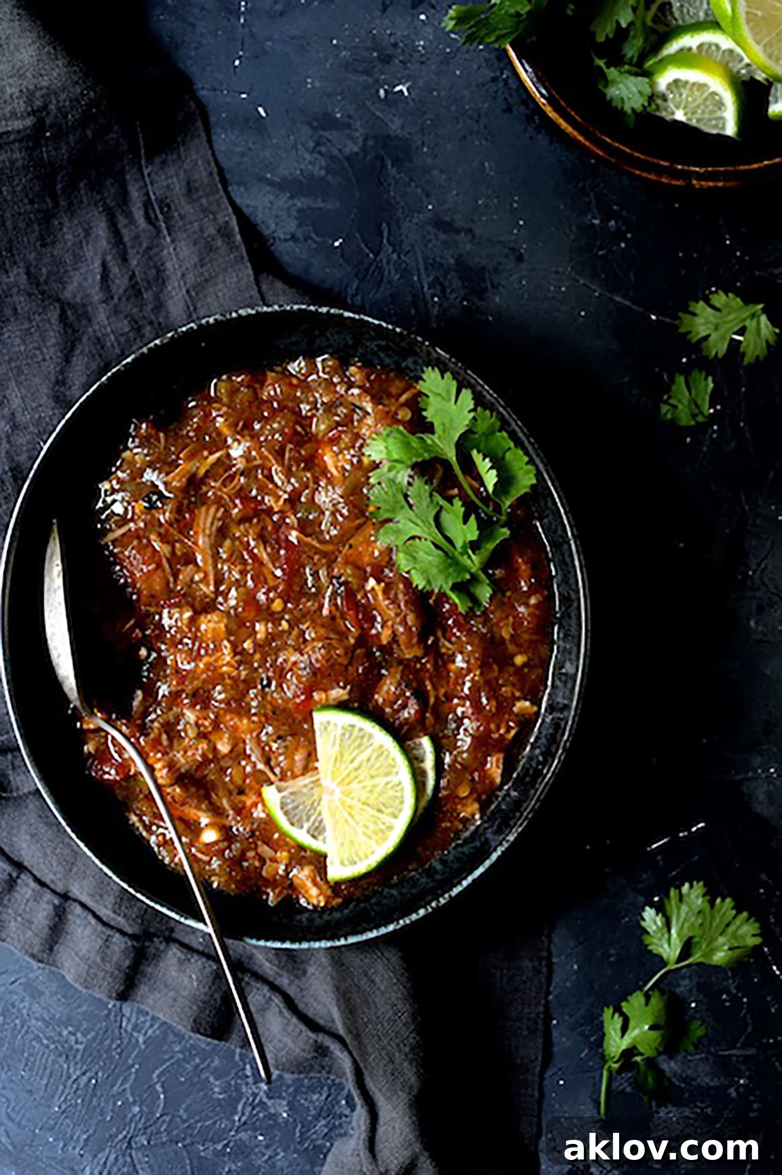 One serving of Slow Cooker Colorado Green Chili in a black bowl on a dark background, garnished with fresh lime wedges and cilantro sprigs. The chili is rich and inviting.