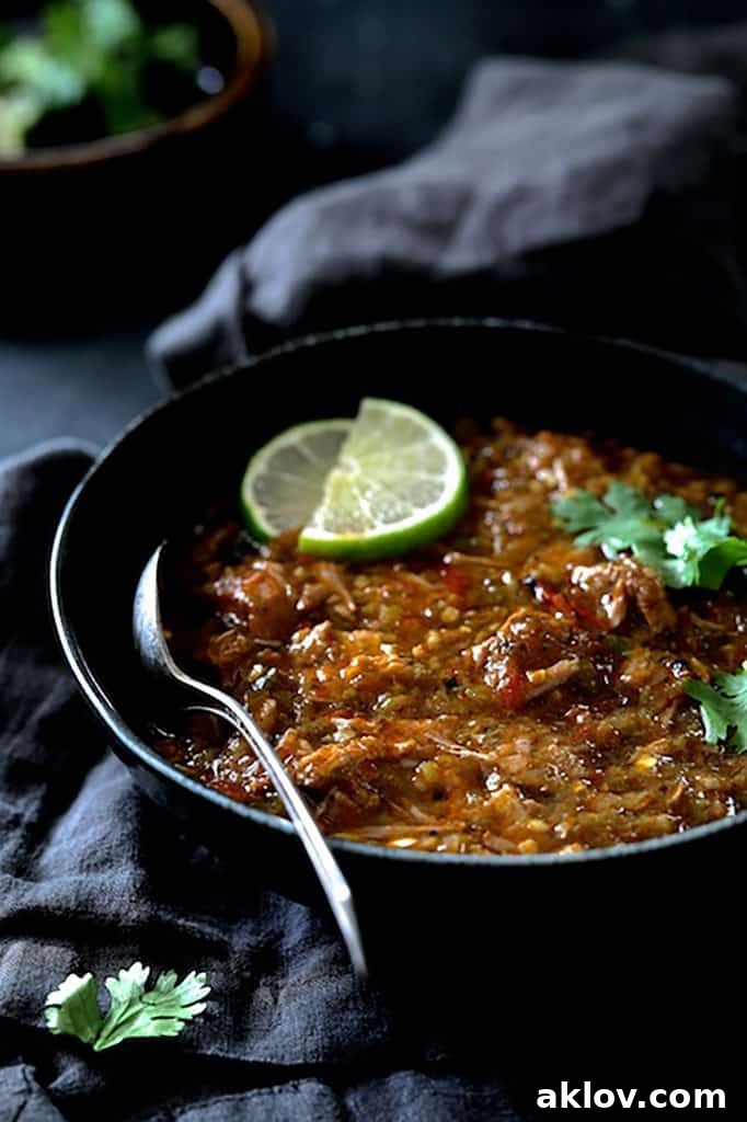 A generous serving of Slow Cooker Colorado Green Chili in a black bowl, resting on a gray napkin. It is garnished with fresh cilantro sprigs and bright lime wedges.