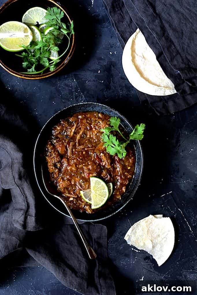 A photo showcasing one serving of Slow Cooker Colorado Green Chili in a black bowl against a dark background, accompanied by fresh lime wedges, cilantro sprigs, and corn tortillas, ready to be enjoyed.